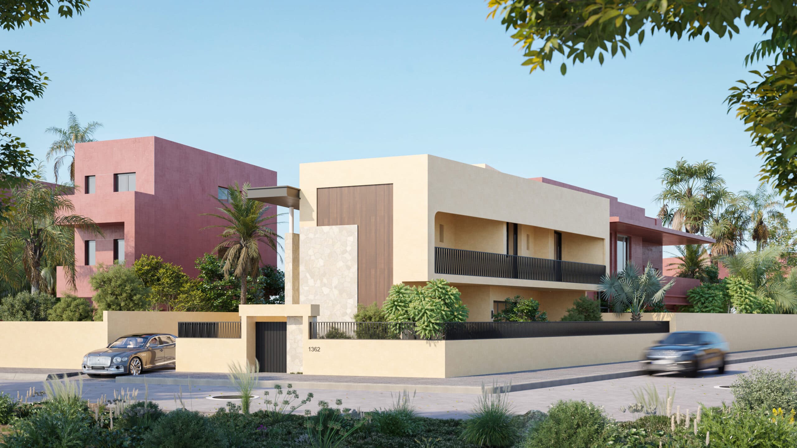 Maison moderne à deux étages avec une façade beige et brune, entourée d'une végétation luxuriante. Une voiture est garée à gauche et une autre roule à droite. Des bâtiments roses et des palmiers sont visibles à l'arrière-plan sous un ciel clair.